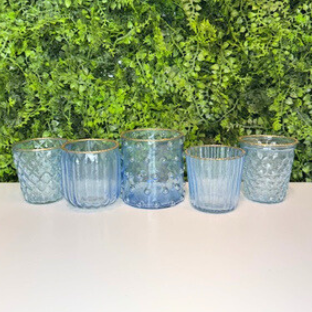 Set of five blue textured glass votives with gold rims on a white surface with green foliage in the background.