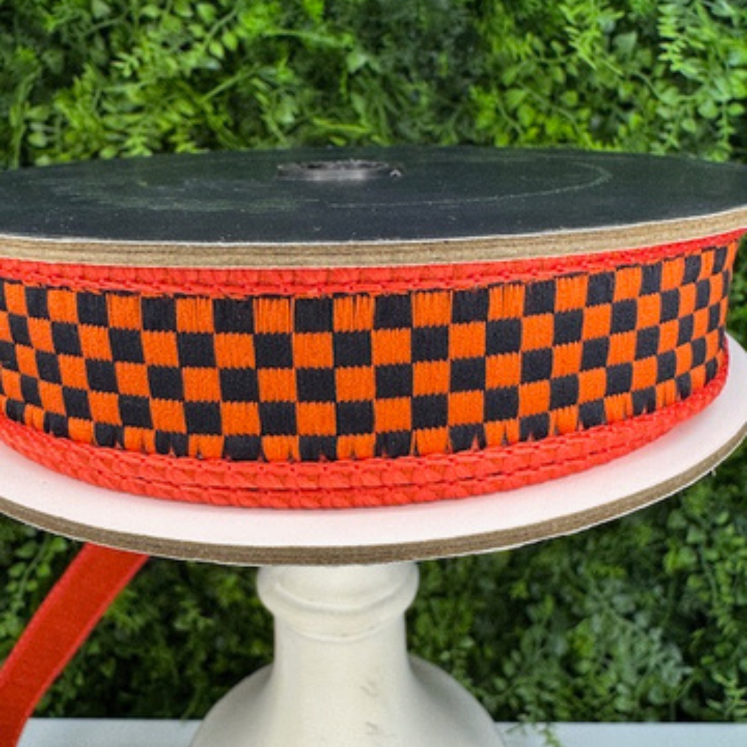 Spool of orange and black Farrisilk checkered ribbon on a white stand with a green foliage background