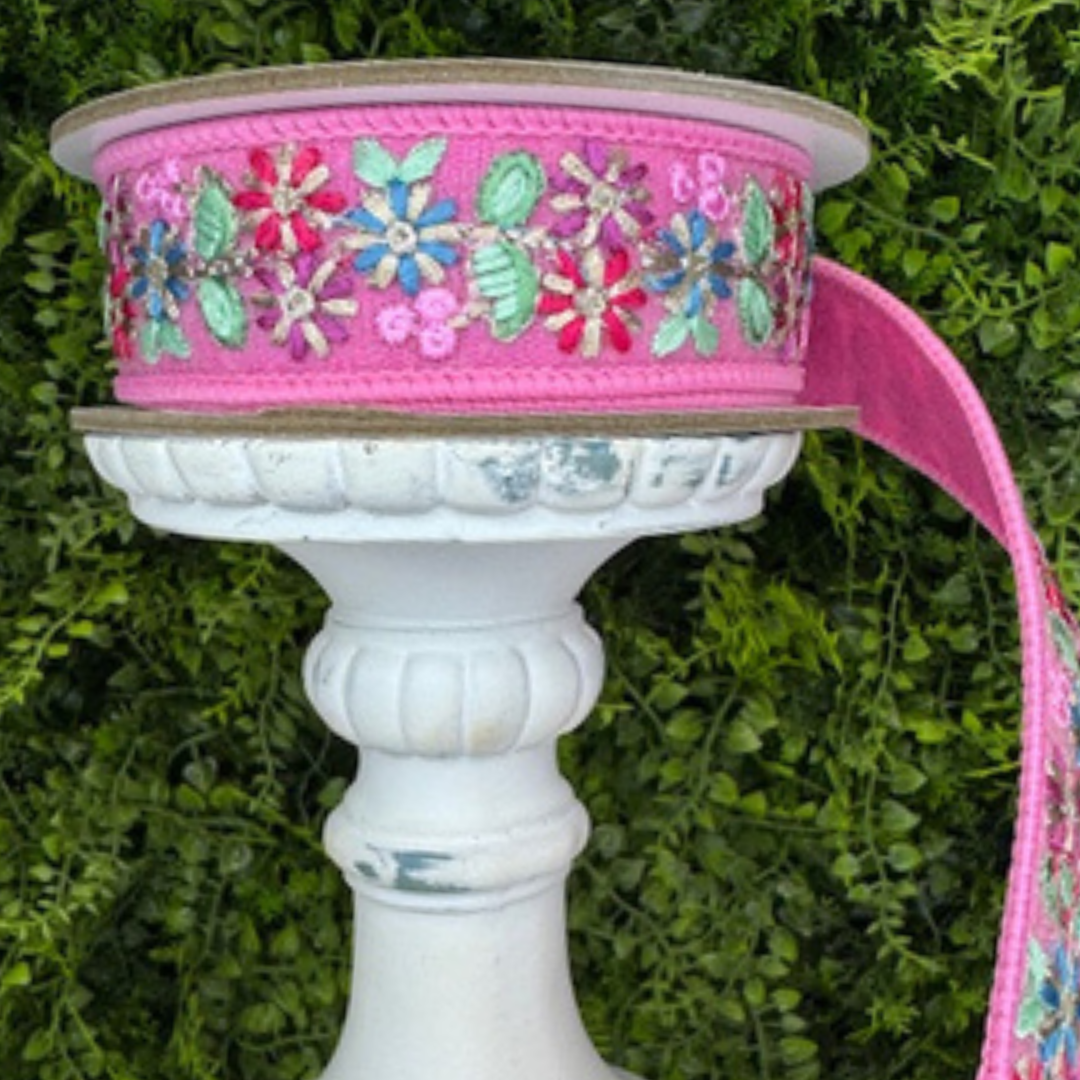 Decorative ribbon spool with floral embroidery on a white pedestal against a green leafy background
