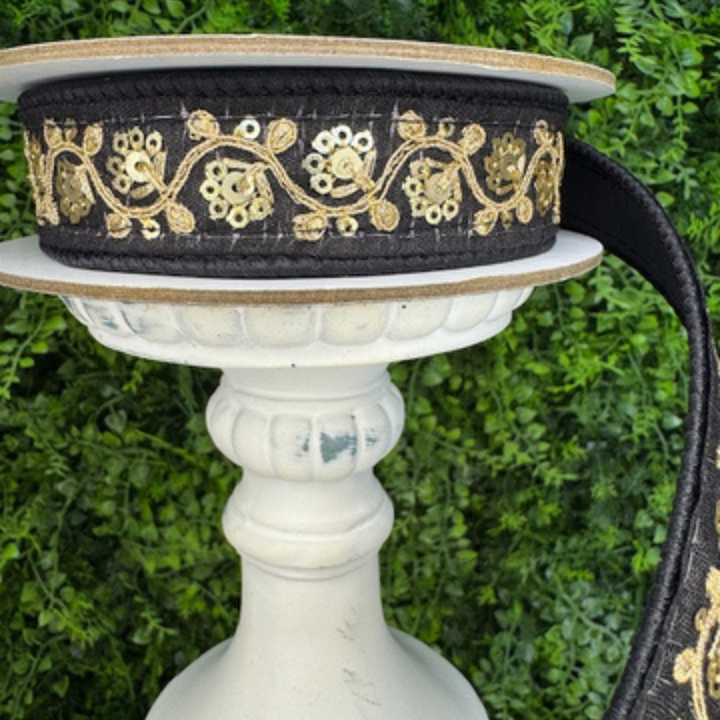 Decorative Farrisilk black ribbon with gold patterns on a white pedestal against a green foliage background